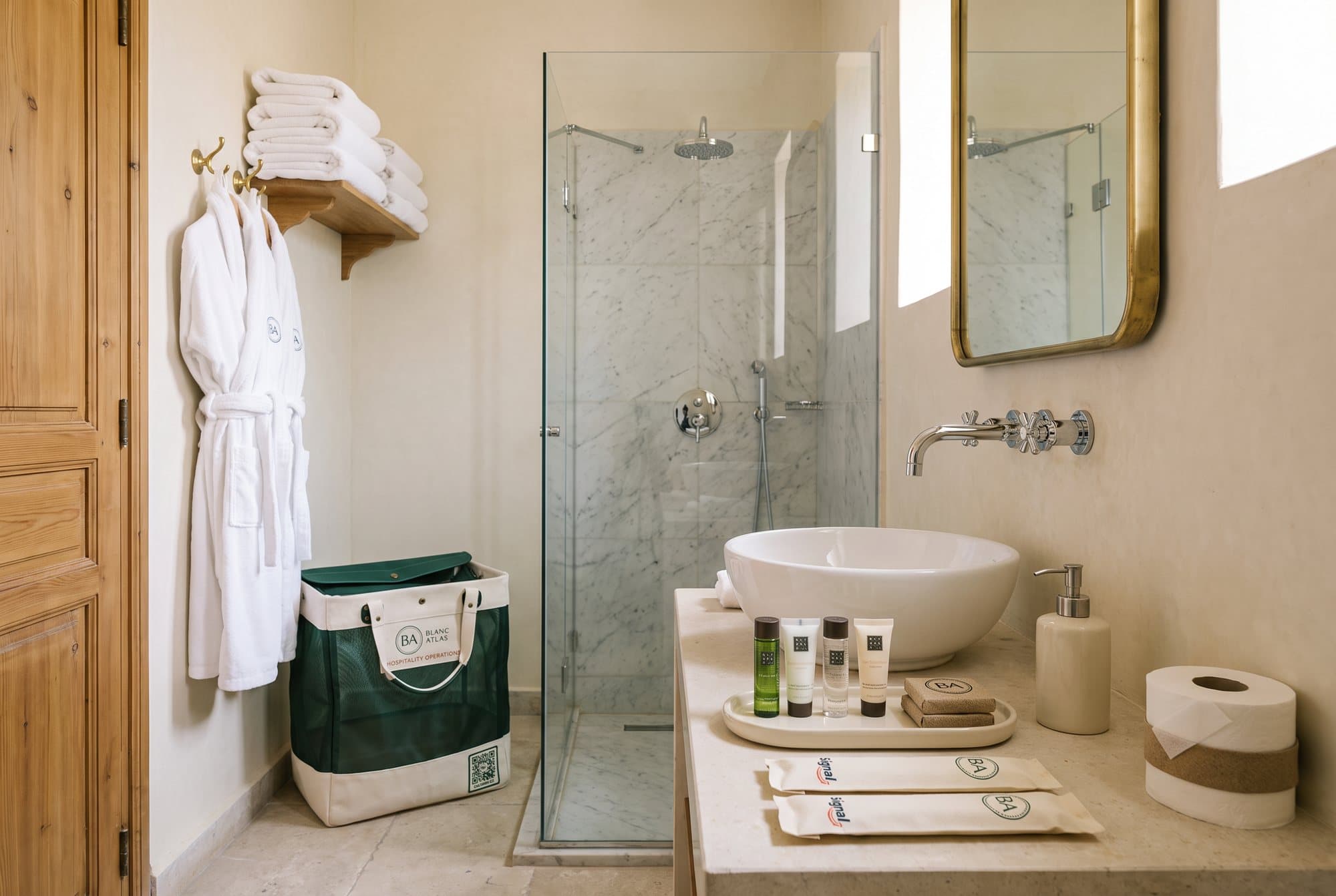 Salle de bain prête : peignoirs et serviettes blanches, douche en marbre impeccable, minis Rituals et kits Signal sur le plan vasque, panier Blanc Atlas vide